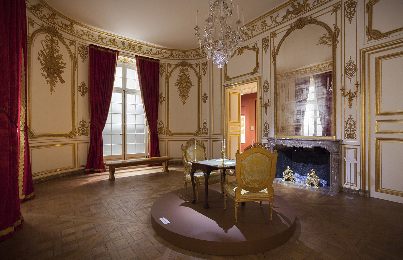 Photograph of an 18th century period room with gilt gold ornamentation, a crystal chandelier, red draperies, and a marble fireplace.